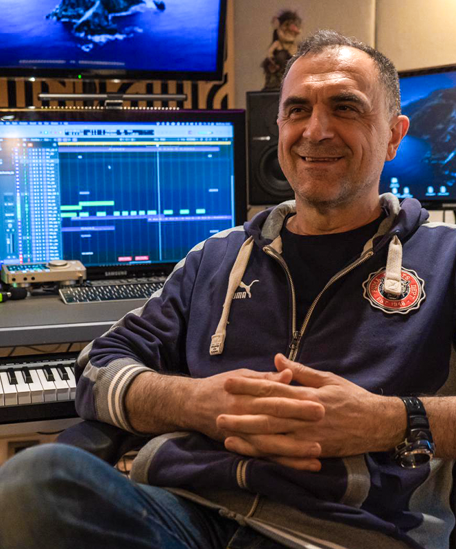 Raniero Gaspari seduto nel suo studio di registrazione, sorridente, con uno schermo di editing musicale alle spalle, tastiera e strumenti musicali in vista.